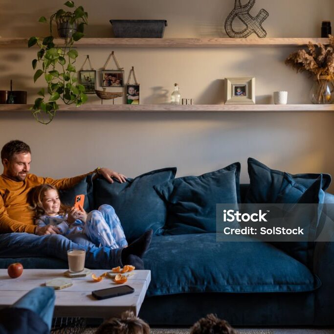 A full shot of a father lying down on the sofa with his daughter lying next to him. The girl is holding a mobile phone with her father looking over her shoulder.